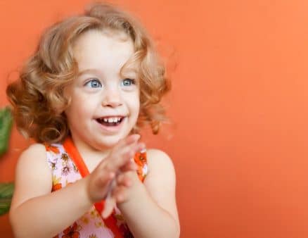Portrait of a beautiful small girl with gorgeous blue eyes and blonde curly hair clapping her hands on an orange background