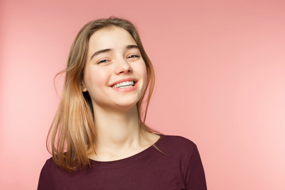 A person with light brown hair smiles brightly against a soft pink background, wearing a burgundy top. The image conveys a cheerful mood.