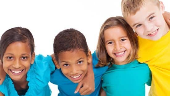 Four smiling children wearing colorful shirts stand close together against a white background, demonstrating friendship and joy.